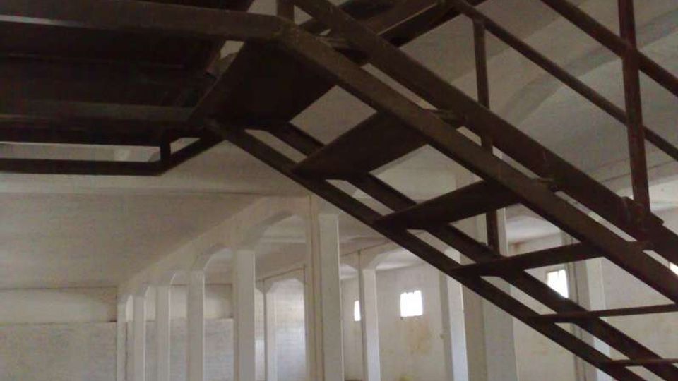 Interno di un grande capannone industriale vuoto con file di pilastri bianchi, pavimento in cemento e piccole finestre alte sulle pareti. In primo piano una scala in metallo con ringhiere attraversa lo spazio in diagonale verso un livello superiore.