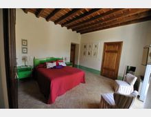 Camera da letto in stile rustico con soffitto in legno a travi a vista, pavimento in piastrelle e pareti bianche con fascia verde. Al centro c’è un letto matrimoniale con copriletto rosso e testiera verde, affiancato da comodini coordinati; sulla destra si vedono due poltroncine a righe e una porta in legno.