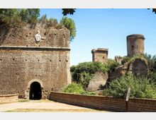 Rovine di una fortificazione in pietra con un grande muro e un arco d’ingresso, affiancata da una passerella e vegetazione fitta. Sullo sfondo si vedono torri e bastioni in pietra sotto un cielo sereno.