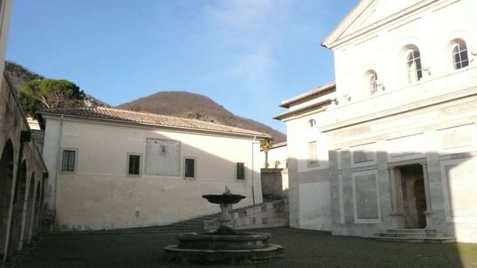 Piazza lastricata in pietra con una fontana circolare al centro, circondata da edifici chiari con finestre e tetti in tegole. Sullo sfondo si vede una collina boscosa sotto un cielo sereno.