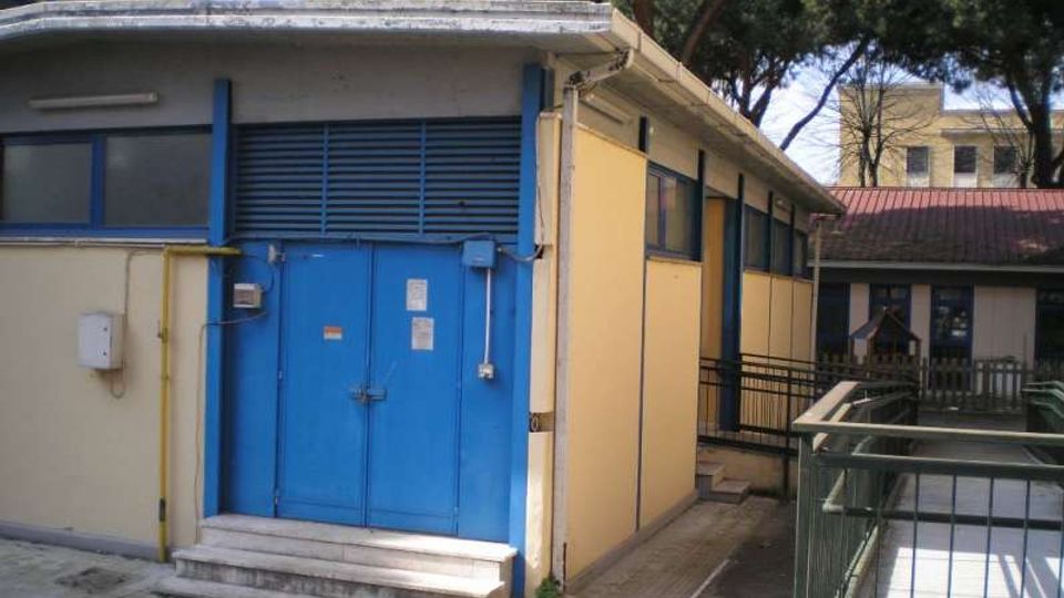 Edificio basso di servizio con facciata gialla e grandi porte metalliche blu, con piccola scalinata d’ingresso e finestre laterali. L’area esterna è pavimentata con una rampa e ringhiere, circondata da alberi e altri edifici sullo sfondo.