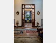 Interno di un salone elegante con pavimento in legno a spina di pesce e tappeti persiani, che conduce attraverso una grande porta ad arco in legno e vetro verso un secondo ambiente con vetrate decorative e una pianta in vaso al centro. Sulle pareti chiare sono presenti piccoli tavolini e ritratti ovali incorniciati.