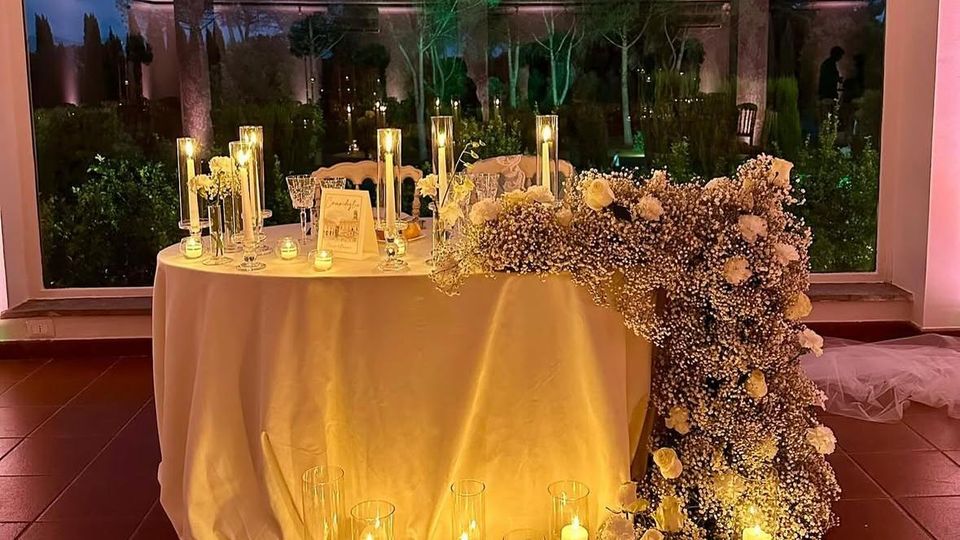 Tavolo rotondo con tovaglia bianca allestito con candele in cilindri di vetro e decorazioni floreali, con un’ampia composizione di fiori bianchi che scende sul lato fino al pavimento. Sullo sfondo, una grande vetrata affaccia su un giardino con alberi e colonne illuminate al crepuscolo.