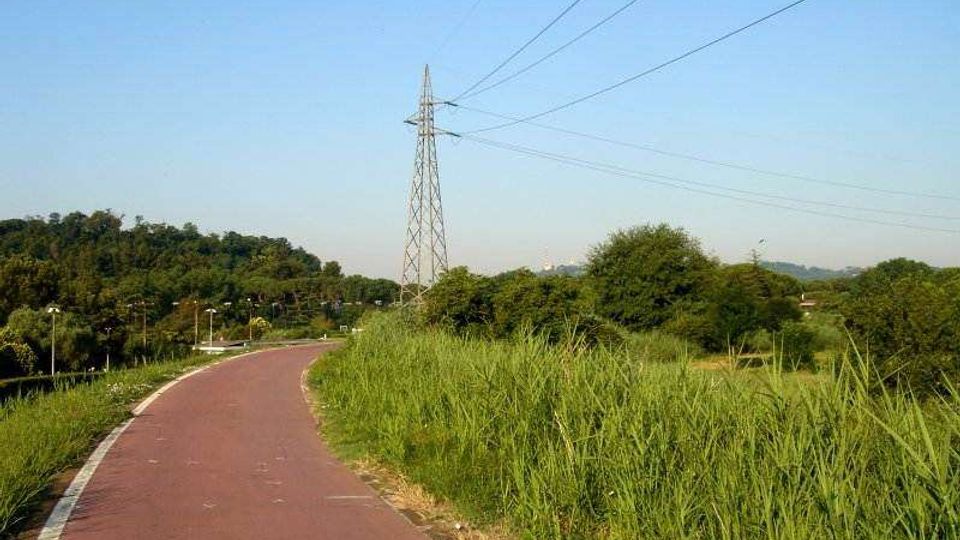 Pista ciclopedonale asfaltata color rosso che curva attraverso un’area verde con erba alta e alberi. Sullo sfondo si vede un traliccio dell’alta tensione con cavi elettrici che attraversano il cielo sopra le colline.