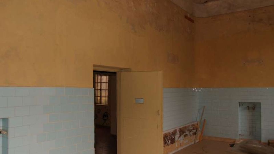 Interno di una stanza spoglia con pareti superiori intonacate color ocra e fascia inferiore rivestita di piastrelle azzurre, con segni di deterioramento e umidità. Al centro c’è una porta aperta che conduce a un ambiente più buio, con pavimento in piastrelle e una finestra con grata visibile sullo sfondo.