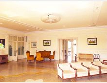 Ampia sala interna in stile classico con pareti chiare decorate da cornici, lampadario centrale e pavimento in graniglia con motivi. Sono presenti divani e poltrone disposte come area lounge, quadri alle pareti e grandi porte vetrate che collegano ad altre stanze.