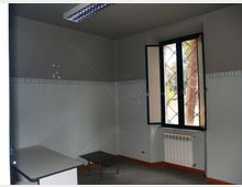 Interno di una piccola stanza d’ufficio con pareti grigie e una fascia decorativa, illuminazione a neon a soffitto e pavimento grigio. Sulla destra c’è una finestra a battenti con inferriata e radiatore sotto, mentre nell’angolo si vedono una scrivania e un tavolino basso.