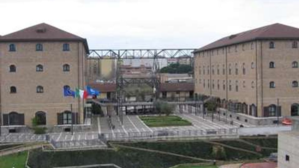 Cortile urbano tra due grandi edifici in mattoni con tetti a falde, collegati da una passerella metallica sopraelevata. In primo piano si vedono aiuole geometriche, camminamenti e un’area verde, con bandiere vicino all’ingresso.