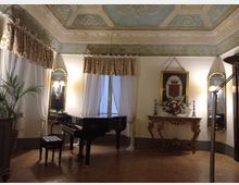 Salone interno in stile classico con soffitto affrescato e cornici decorative, pavimento in cotto e grandi finestre con tende bianche e mantovane floreali. Al centro un pianoforte a coda nero, accanto a una consolle intagliata con composizione floreale, specchi con appliques e un quadro con stemma sulla parete.