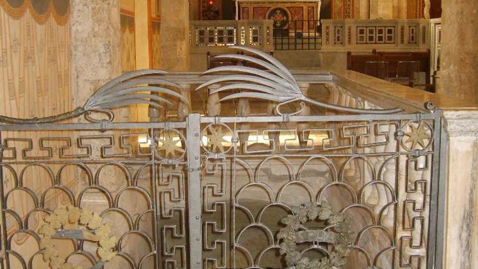Interno di una chiesa con colonne in pietra e pareti affrescate; in primo piano una cancellata in ferro battuto decorata delimita un’area ribassata. Sullo sfondo si vede l’altare con tabernacolo e un’abside con affreschi e finestra centrale.