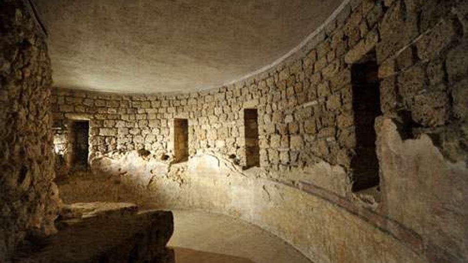 Interno di una struttura sotterranea in pietra con pareti curve in blocchi squadrati e soffitto a volta. Lungo il muro si aprono piccole nicchie rettangolari e il pavimento in terra segue l’andamento semicircolare dell’ambiente.