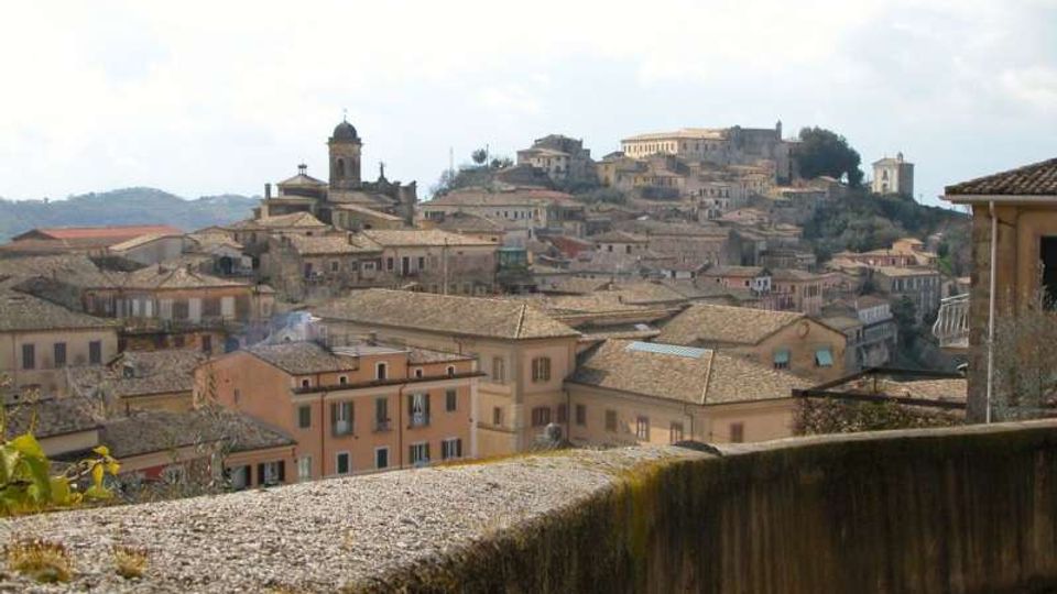 Veduta panoramica di un borgo collinare con case dai tetti in coppi e edifici storici addossati, tra cui una torre campanaria e una chiesa. In primo piano si vede un muretto in pietra, con il centro abitato che si estende sulle pendici sotto un cielo parzialmente nuvoloso.