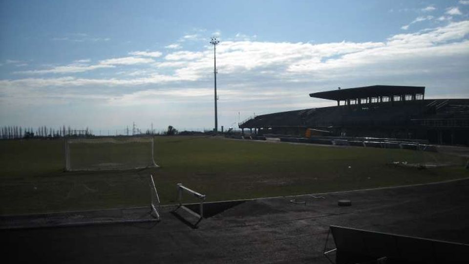 Campo da calcio all’aperto con pista perimetrale e piccole porte, illuminato da un alto palo faro. Sul lato destro si vede una tribuna coperta e bassa, con il cielo aperto sullo sfondo.