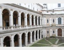 Cortile interno in pietra con portici su due livelli, scanditi da archi e colonne, e balaustra al piano superiore. Sullo sfondo si vede una facciata curva con finestre ad arco e un grande portone, affacciata su un pavimento a motivi geometrici.