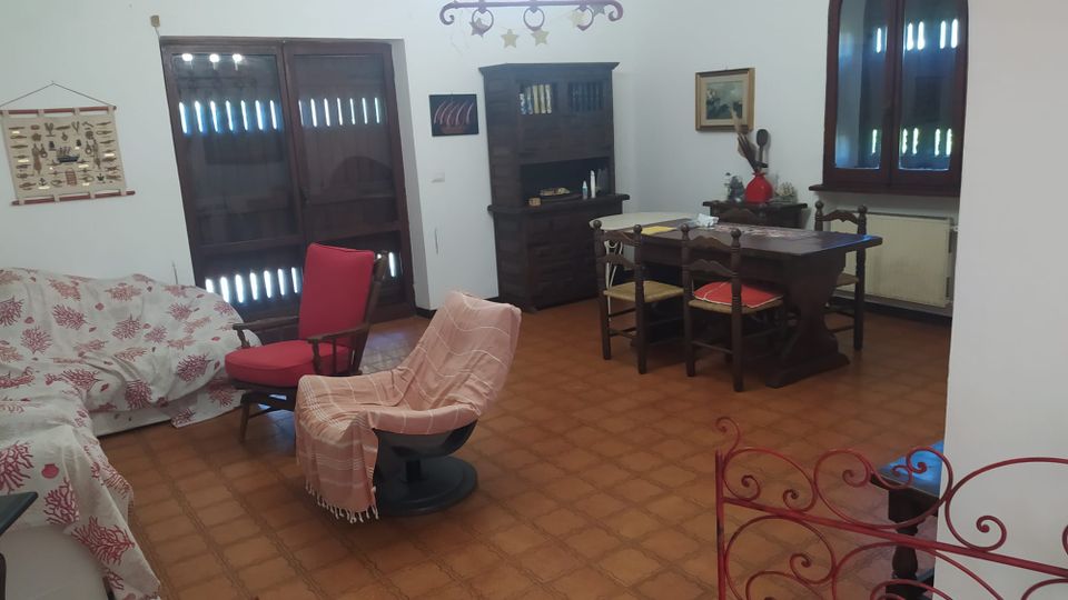 Soggiorno-sala da pranzo con pavimento in cotto, tavolo in legno con sedie e credenza scura contro la parete. Sono presenti un divano con copridivano decorato, poltrone, lampadario a soffitto e due grandi aperture con infissi in legno (porta-finestra e finestra ad arco).