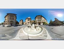 Grande piazza urbana con una fontana monumentale in pietra al centro di una rotonda, circondata da ampie strade asfaltate. Intorno si vedono palazzi storici di più piani con facciate decorate e balconi, sotto un cielo sereno e luminoso.