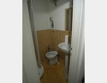 Piccolo bagno con pareti e pavimento in mosaico giallo, con wc e lavabo a colonna accanto a uno specchio. A sinistra si vede una cabina doccia semicircolare con ante in vetro.