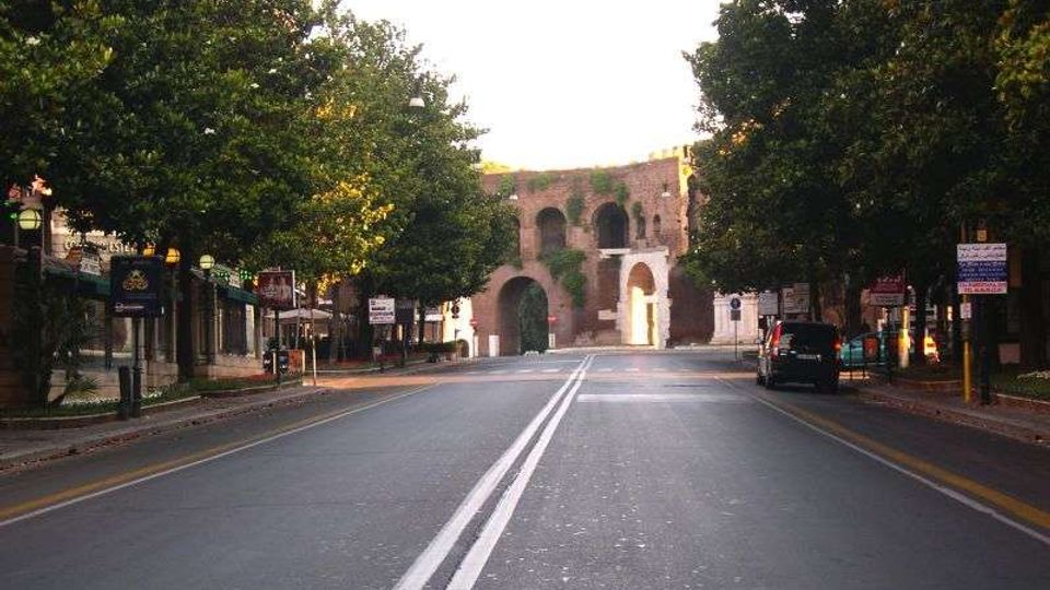 Strada urbana a due corsie con doppia linea continua centrale, fiancheggiata da alberi e edifici bassi. In fondo si vede una grande struttura in mattoni con archi e aperture, simile a una porta o resti di fortificazione.