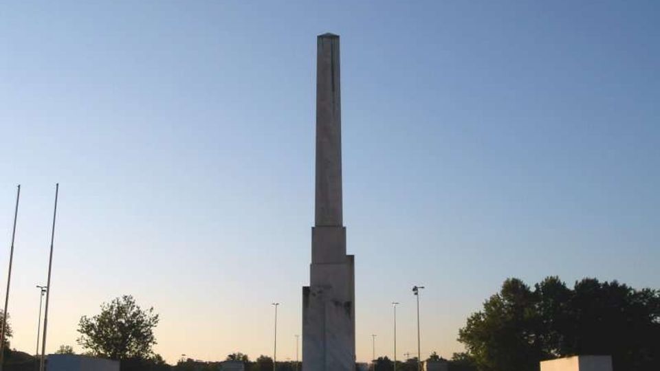 Obelisco in pietra su alto basamento al centro di una rotonda o piazza, circondato da piccoli arbusti e blocchi squadrati. Sullo sfondo si vedono alberi, alcuni lampioni stradali e un cielo sereno al tramonto.
