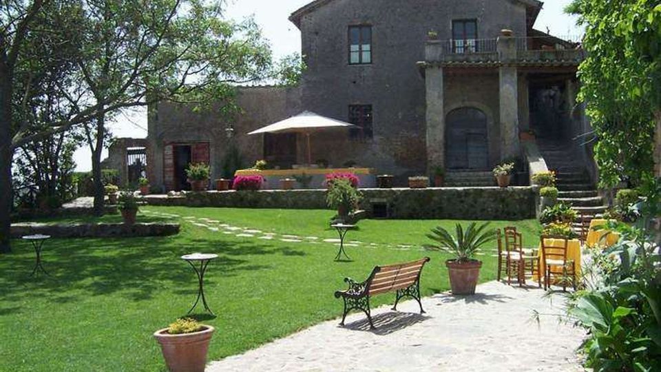Cortile-giardino con ampio prato curato, vialetto in pietra e vasi di piante, con panchina e piccoli tavolini da esterno. Sullo sfondo si vede un edificio rurale in pietra a due piani con terrazza, scala esterna e un patio con ombrellone, circondato da alberi.