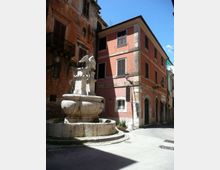 Piazzetta in un centro storico con una fontana in pietra al centro, sormontata da una scultura alata. Intorno si vedono edifici a più piani con facciate in intonaco, finestre con persiane e lampioni a muro lungo una strada stretta.
