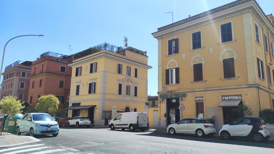 Incrocio urbano con edifici residenziali di tre-quattro piani in tinte pastello, persiane scure e negozi al piano terra con insegne e tende. In primo piano si vedono strisce pedonali, auto parcheggiate e cassonetti lungo il marciapiede sotto un cielo sereno.