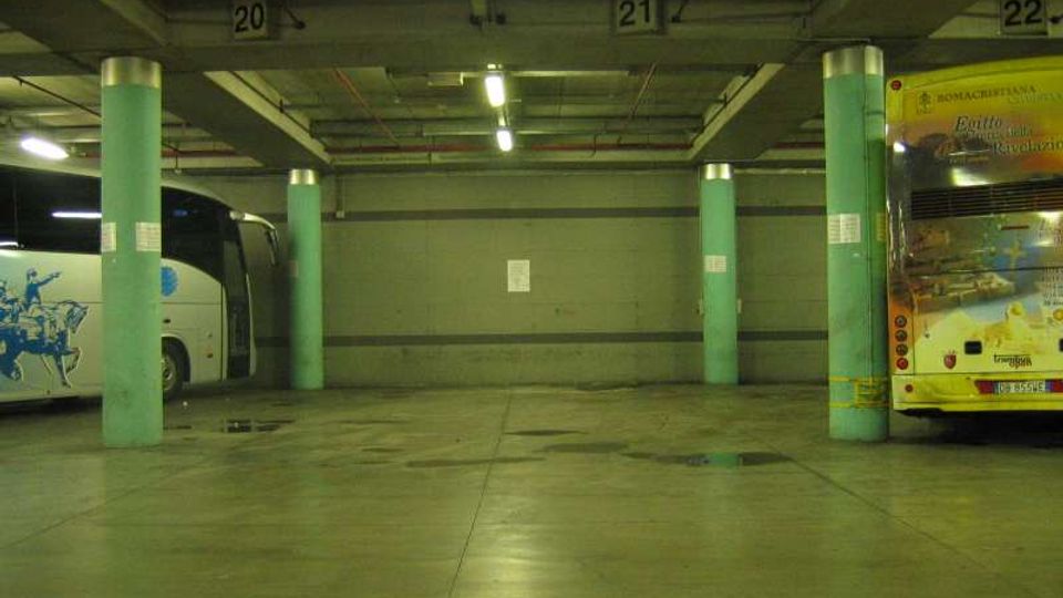 Interno di un’autorimessa o terminal bus sotterraneo con pavimento in cemento lucido, colonne cilindriche verdi e illuminazione a neon sul soffitto. Si vedono due autobus parcheggiati ai lati e numeri di stallo (20, 21, 22) segnati sulle travi superiori.