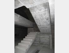 Interno in cemento grezzo di un edificio o bunker sotterraneo, con una scala in calcestruzzo che sale verso un solaio inclinato. Travi e pareti massicce in cemento a vista creano un ambiente angusto e poco illuminato.