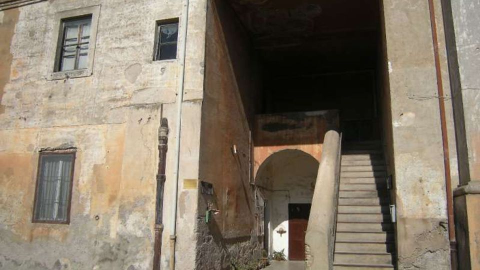 Facciata di un edificio antico con intonaco consumato e finestre rettangolari, affacciata su una strada. A destra si apre un grande passaggio coperto con una scalinata in pietra che sale lungo il muro e conduce a un cortile interno con arco e portone.