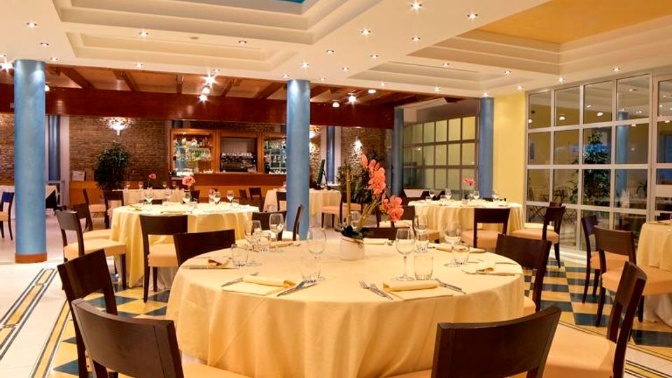 Sala ristorante elegante con tavoli rotondi apparecchiati con tovaglie chiare e sedie scure, su pavimento a motivi geometrici blu e gialli. Soffitto a livelli con riquadro centrale azzurro e faretti, colonne blu, pareti vetrate laterali e area bar sul fondo.