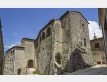 Antico edificio in pietra con facciata irregolare, grandi archi murati e finestre ad arco, affacciato su una piccola piazza lastricata con scalinate e muretti. Sullo sfondo si vedono altre case in pietra e un cielo azzurro con alcune nuvole.