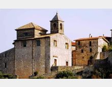 Edifici storici in pietra su un pendio, con una chiesa dal campanile quadrato e tetto a piramide in primo piano. Intorno si vedono case in muratura con tetti in coppi e un muretto di contenimento in pietra sotto un cielo limpido.