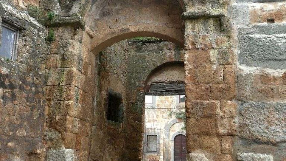 Antico passaggio ad arco in pietra tra muri spessi e irregolari, con tracce di vegetazione tra le fughe. Il pavimento è in lastre di pietra e l’arco conduce a un piccolo cortile con un edificio in muratura e una porta sullo sfondo.