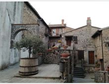 Piccola piazza in un borgo con case in pietra, archi e scale che collegano livelli diversi. Al centro ci sono grandi fioriere e un ulivo in una botte, con muri in pietra e porte in legno sullo sfondo.