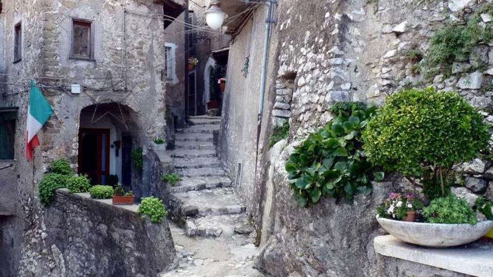 Vicolo stretto in un borgo in pietra con ripida scalinata di gradini irregolari tra muri e case in muratura, con lampione sospeso e cavi a vista. Piante in vaso e una piccola fioriera su muretto di pietra, con ingresso ad arco e bandiera italiana su una facciata.