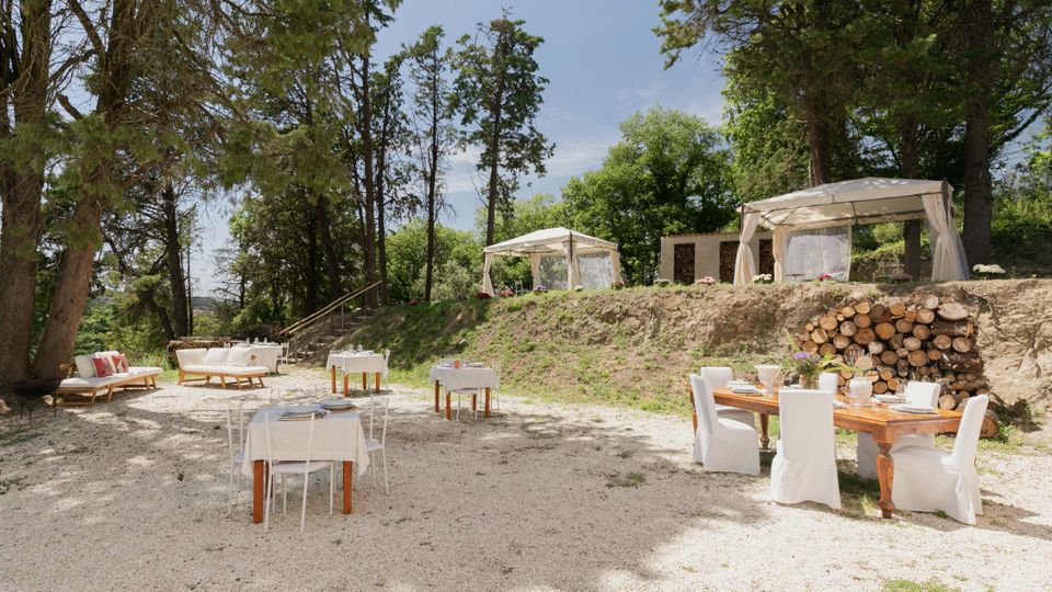 Area esterna attrezzata per ristorazione in un bosco, con tavoli su ghiaia e sedie con coperture bianche sotto alberi alti. Su un terrapieno sullo sfondo ci sono due gazebo bianchi e una catasta di legna accanto a una parete in pietra.