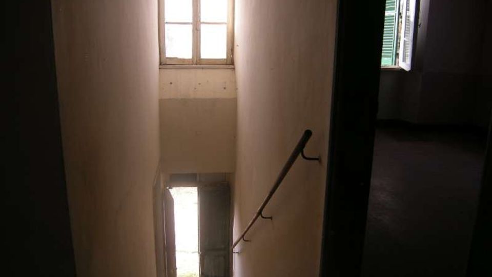 Vano scala interno stretto con pareti chiare, gradini in discesa e corrimano metallico a destra. Una porta aperta al piano inferiore e una finestra alta illuminano lo spazio, con un’altra finestra laterale dotata di persiane verdi.