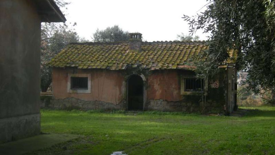Piccola casa rurale in muratura con tetto a due falde coperto di tegole e muschio, con ingresso ad arco e finestre con inferriate. Davanti si estende un prato verde, con alberi ai lati e un altro edificio parzialmente visibile sulla sinistra.