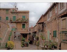 Cortile in un borgo con case in pietra e intonaco, persiane verdi, balconi con fioriere e scale esterne con ringhiere. Pavimentazione in pietra, vasi di piante e piccoli ingressi sotto portici, con cielo nuvoloso sullo sfondo.