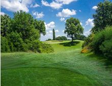 Ampio campo da golf con prato curato e lievi ondulazioni, circondato da alberi e cespugli. Sullo sfondo si vedono un green con bunker di sabbia e un alto cipresso sotto un cielo azzurro con nuvole sparse.