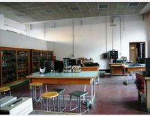 Interno di un laboratorio scolastico di elettronica con banchi da lavoro e sgabelli, strumenti e apparecchiature tecniche appoggiati sui tavoli. Lungo la parete sinistra ci sono armadi vetrati con materiale, soffitto a pannelli e cavi a vista sulle pareti, con una porta sul fondo.