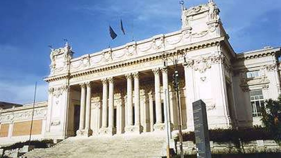 Facciata monumentale di un edificio neoclassico con ampio portico a colonne e lunga scalinata in pietra che conduce all’ingresso. La struttura chiara presenta decorazioni e frontone con bandiere, affacciata su una strada con auto in primo piano e cielo sereno sullo sfondo.