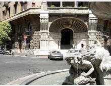 Ingresso di un edificio storico in pietra con grande arco decorato e scalinate, affacciato su una strada urbana. In primo piano si vede una fontana monumentale in pietra con sculture e vasca circolare.