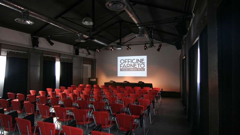 Sala conferenze in stile industriale con soffitto alto nero, travi metalliche e condotti a vista, illuminazione a binari e faretti. File ordinate di sedie rosse rivolte verso uno schermo di proiezione sul fondo con un palco basso.