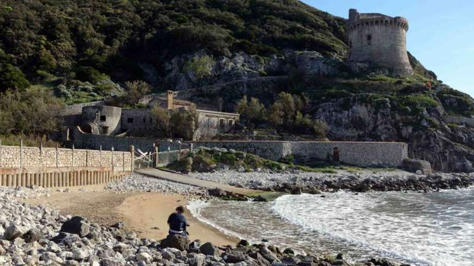 Piccola spiaggia con ciottoli e riva sabbiosa affacciata sul mare, con onde che arrivano a riva e un camminamento costiero protetto da muretti in pietra. Sul pendio roccioso e boscoso retrostante si vede un complesso fortificato con una torre circolare in pietra in cima.