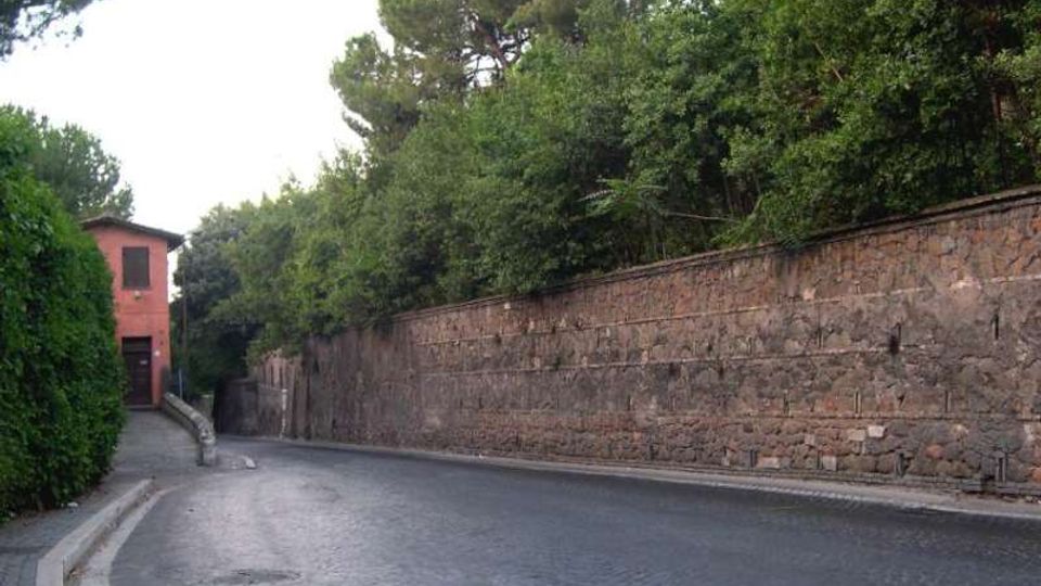 Strada curva in pavé accanto a un lungo muro in pietra sormontato da vegetazione fitta e alberi. Sul lato sinistro si vede un marciapiede che conduce a una piccola casa rosa con ingresso su una stradina laterale.