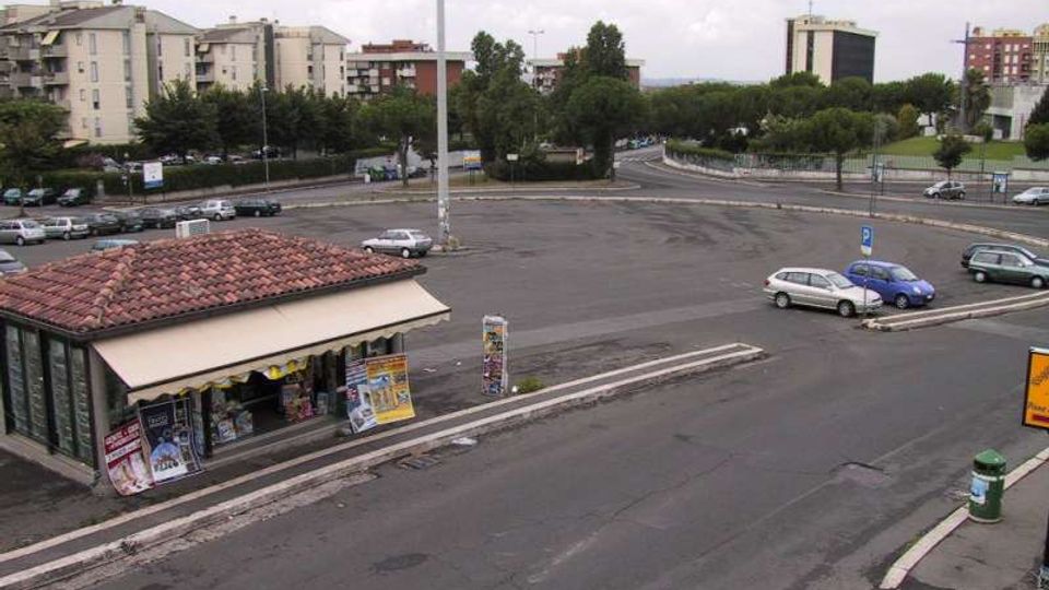 Ampio parcheggio asfaltato con strisce di sosta e un’edicola a un piano con tetto in tegole rosse in primo piano. Sullo sfondo si vedono alberi, strade di raccordo e palazzi residenziali e uffici.