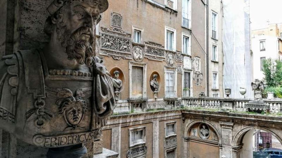 Cortile interno di un palazzo storico in pietra, con facciate decorate da rilievi e nicchie con statue, logge ad arco e una balaustra. In primo piano si vede un grande busto in marmo su piedistallo, affacciato sul cortile.