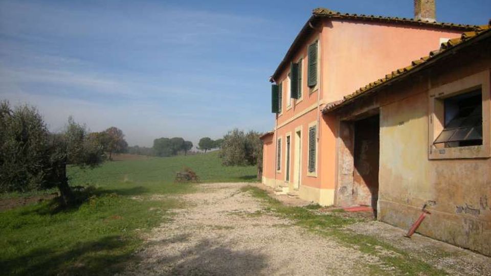 Casa di campagna a due piani intonacata color rosa con persiane verdi e tetto in coppi, affiancata da un annesso più basso con finestra. Davanti si estende un vialetto di ghiaia che conduce a campi verdi con alberi sparsi sotto un cielo sereno.