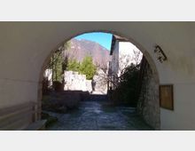Passaggio ad arco in muratura che incornicia un vialetto acciottolato verso un cortile con edifici in pietra e vegetazione. Sullo sfondo si vedono alberi e montagne, con una parete in pietra e una lampada a muro lungo il lato destro.